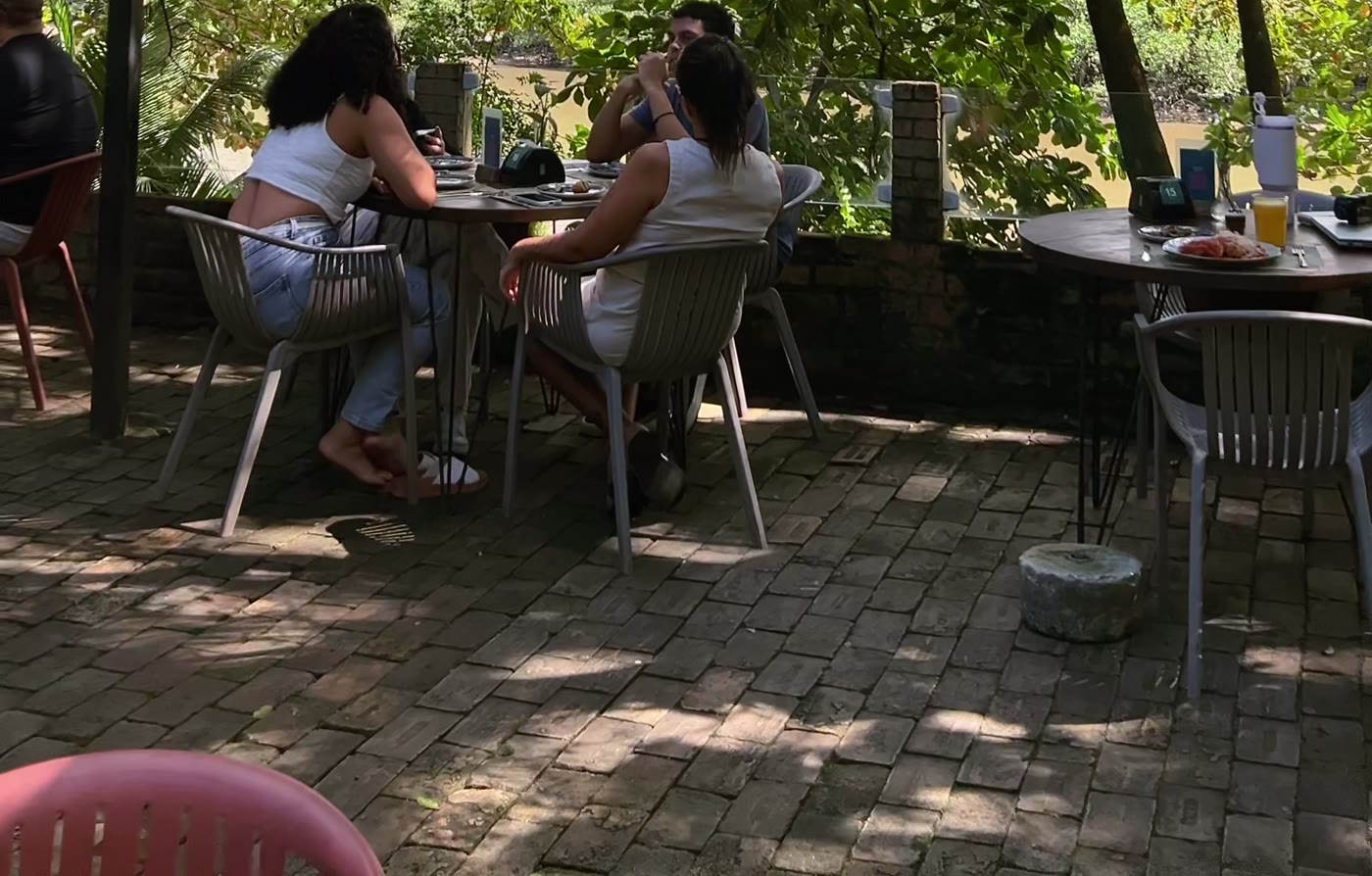 Clientes almoçando nas mesas ao ar livre, no piso de pedra sob a sombra das árvores.